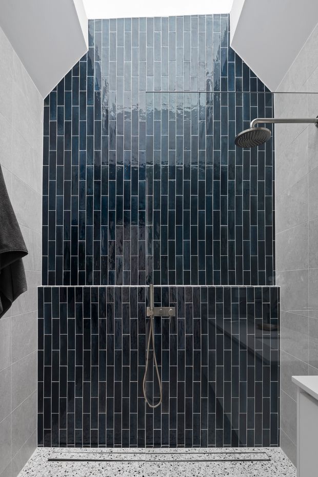 A contemporary walk-in shower with a full-height feature wall clad in glossy dark blue-grey vertical rectangular tiles. Light grey wall tiles flank the shower, with a brushed metal rainfall showerhead and handheld fixture, a clear glass screen, and a terrazzo-like shower floor. The space is compact, clean, and high-contrast.