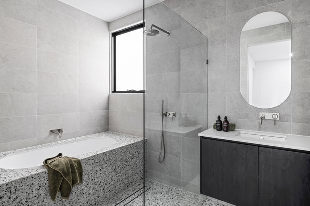 Contemporary bathroom featuring large-format light grey wall tiles, a frameless glass shower enclosure, freestanding-style bath with terrazzo-like speckled surround, dark timber vanity cabinetry, white benchtop, and a rounded-rectangle wall mirror. The space is bright, restrained, and monochromatic with contrasting dark joinery.