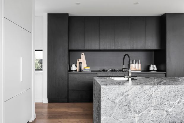Contemporary kitchen with a large waterfall-edge stone island in the foreground, dark full-height cabinetry and a dark splashback wall, integrated cooktop, undermount sink with black gooseneck tap, and timber flooring. White full-height cabinetry frames the space, creating a high-contrast monochromatic scheme with warm wood underfoot.