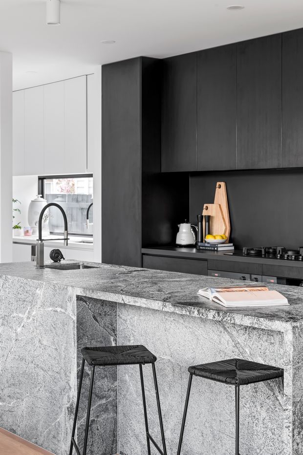 Contemporary kitchen featuring a dramatic dark timber or black fixed cabinetry wall, matching dark splashback, integrated cooktop and black fixtures. A large island is clad in heavily veined grey natural stone with a waterfall edge and two black woven bar stools. White walls and cabinetry, a window above the sink, and minimal black-and-white styling create a refined, monochromatic interior.