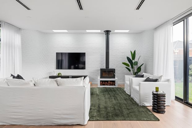 Bright contemporary living room with white-painted brick walls, a central black wood-burning stove and flue, wall-mounted television, white slipcovered sofas and armchairs, a dark green rug, pale timber flooring, sheer curtains, and expansive glazed doors opening to the outdoors.