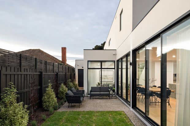 A narrow landscaped courtyard beside a contemporary white multi-level residence. Full-height black-framed glazing opens the interior dining and living areas toward a gravel-paved outdoor seating zone, with a dark timber privacy fence, planting, lawn, and a simple flat-roof form.
