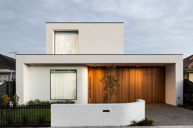Street-facing view of a contemporary two-storey residence with a minimalist flat-roofed form, white rendered and painted brick walls, dark-framed windows, a broad covered entry, and a warm timber-clad garage or entry door. A curved white brick front wall and simple planted garden sit behind a black metal fence.