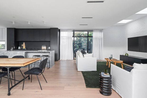 Open-plan contemporary kitchen, dining and living area with light timber flooring, dark charcoal cabinetry, a stone island bench, black-framed glazing, white-painted brick walls and a large wall-mounted television. The space is furnished with a timber dining table, black chairs, white sofas and a dark green rug, creating a clean, modern monochrome interior with natural wood accents.