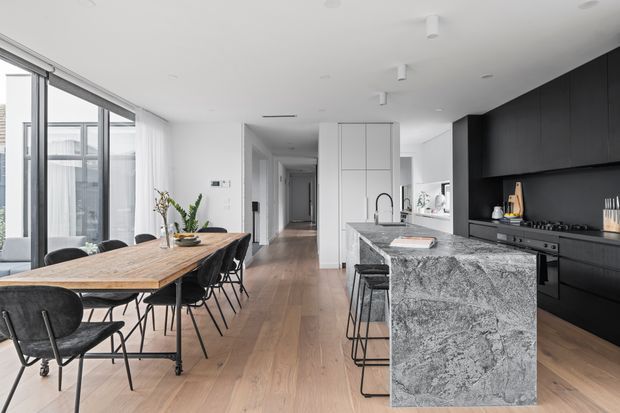 A bright, open-plan contemporary kitchen and dining area. The space features a long natural timber dining table with black chairs, extensive floor-to-ceiling glazing on the left, warm timber flooring, a large grey-veined stone island with bar seating, matte black kitchen cabinetry, integrated appliances, and a white partition wall leading toward a hallway.