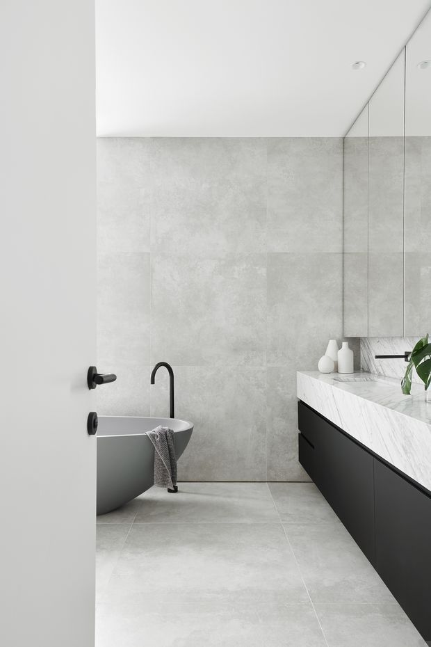 A minimalist contemporary bathroom viewed through a partially open door. The room features large pale grey concrete-look wall and floor tiles, a freestanding grey bathtub with matte black wall-mounted tapware, a long black floating vanity with a white marble-look benchtop, mirrored wall cabinets, and subtle recessed ceiling lighting. The restrained monochrome palette creates a clean, refined atmosphere.