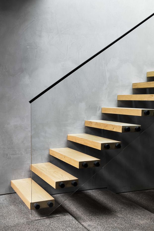 Interior close-up of a modern floating staircase with thick light timber treads, black concealed or metal supports, and a slim black handrail. The stair rises along a textured grey plaster wall, with a transparent glass balustrade and dark speckled flooring below. The composition highlights the cantilevered construction and minimalist detailing.