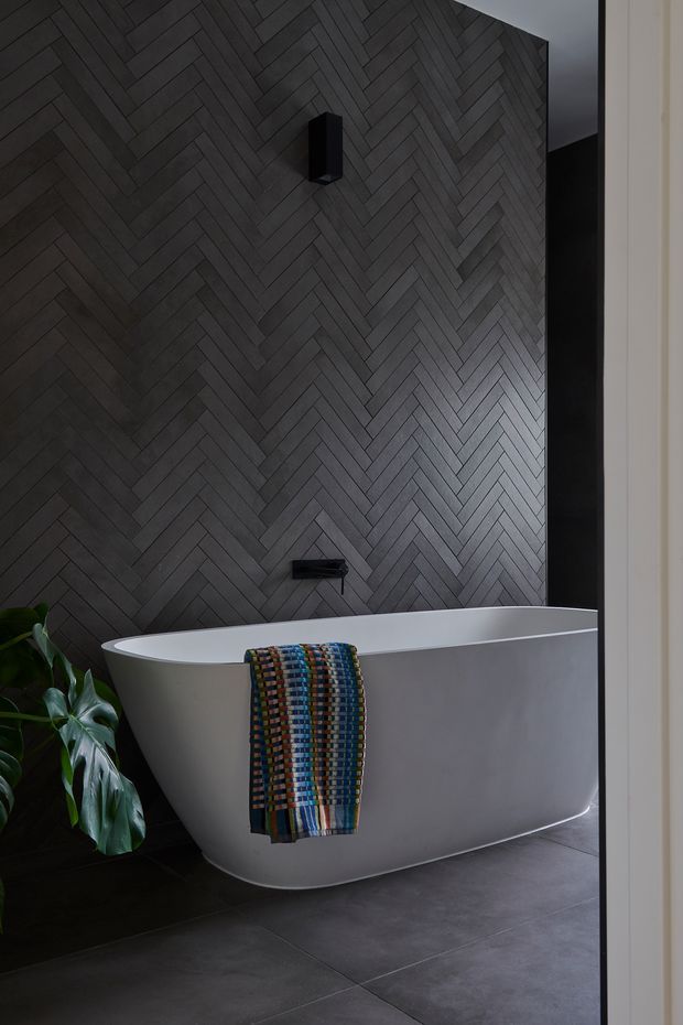 A moody contemporary bathroom featuring a freestanding white bathtub against a dark grey herringbone-tiled feature wall. Matte black wall-mounted tapware and a black wall light provide contrast, while dark floor tiles and a leafy plant reinforce the refined, dramatic atmosphere. A colourful woven towel is draped over the bath.