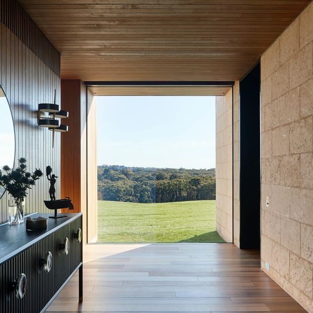 A refined interior passage or entry space framed by a large open glazed doorway overlooking a broad lawn and tree-covered landscape. The room features a warm timber-lined ceiling and floor, a ribbed dark built-in console with sculptural decor, a dark vertical-panel wall, and a textured pale stone masonry wall. Strong natural light and the expansive opening create a calm, indoor-outdoor connection.