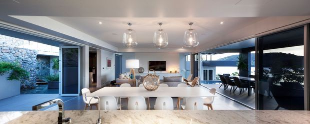A bright open-plan contemporary kitchen and dining/living area viewed from behind a stone island benchtop. The kitchen features a large pale stone island with a stainless-steel sink and tap, white bar stools, white cabinetry, and three pendant lights. Full-height sliding glass doors open to a courtyard on the left and an outdoor or covered dining area on the right, with a lounge and fireplace visible beyond the island.