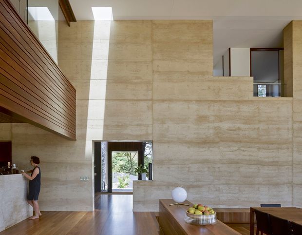 Double-height contemporary interior with a warm, monolithic stone or concrete feature wall, clerestory-style opening, skylight, and timber-lined mezzanine or balcony. The open-plan space includes a timber dining table in the foreground, a small kitchen or service area to the left, dark-framed glazing at the entry, and polished timber flooring. Natural light and strong horizontal and vertical planes create a refined architectural character.