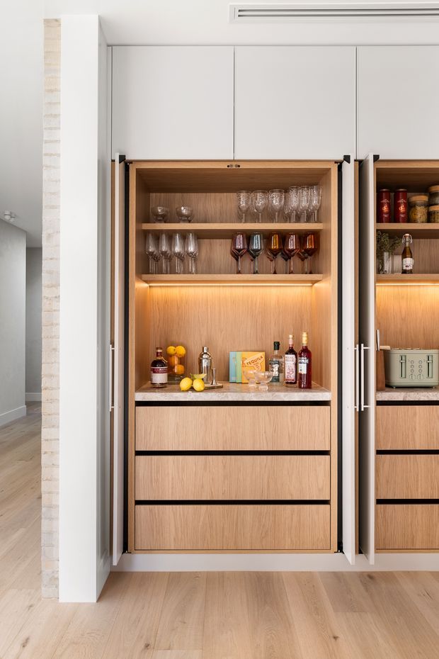 A built-in home bar concealed within tall cabinetry, with warm timber interiors, illuminated open shelving displaying wine and cocktail glasses, bottles and bar accessories, and lower drawers. The cabinetry has white outer doors and slim dark hardware, set against a light timber floor and pale walls.