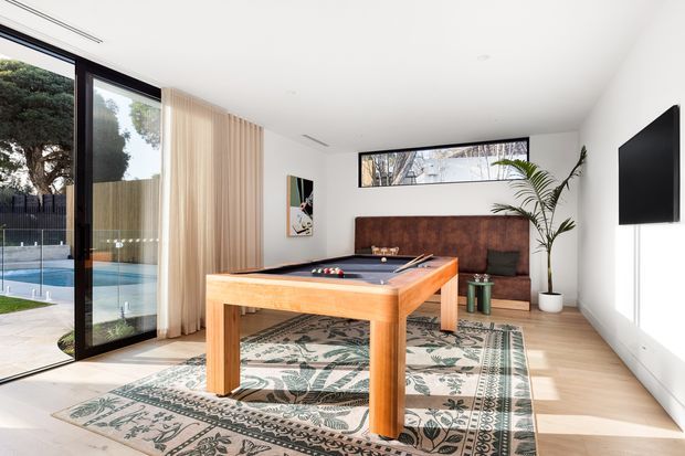 A bright contemporary recreation room featuring a full-size wooden pool table, patterned area rug, built-in dark timber bench seating, a wall-mounted television, and a potted palm. Floor-to-ceiling glazing and sliding doors open to an outdoor swimming pool, with pale timber flooring and white walls creating an airy setting.