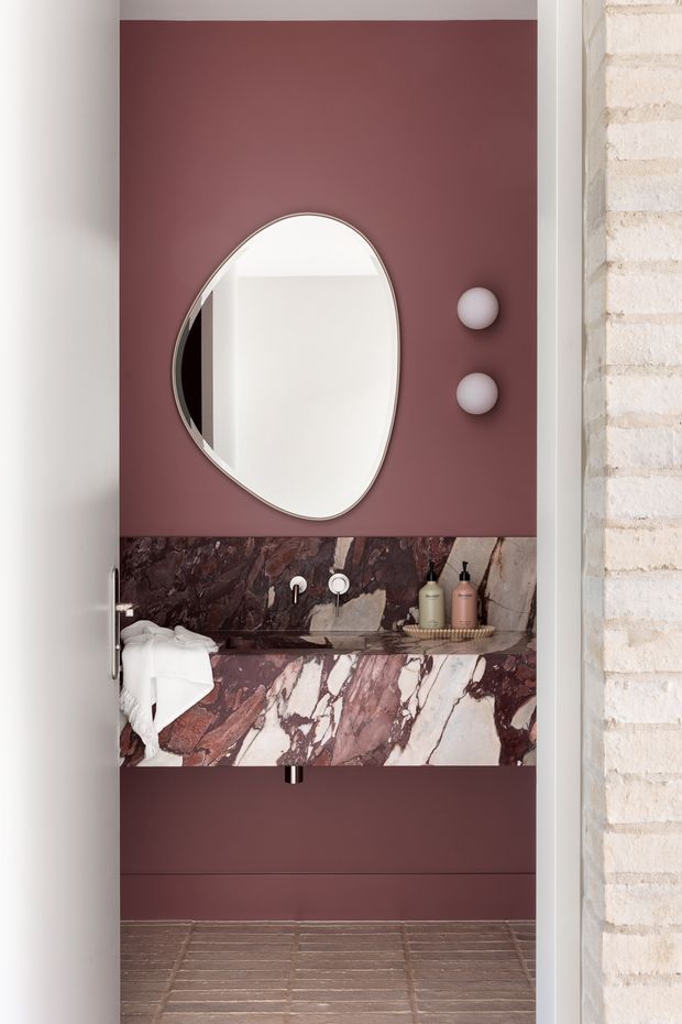 A compact, design-forward bathroom viewed through an open doorway. The room features a deep burgundy painted wall, a large organic oval mirror, two round wall lights, a dramatic pink-and-burgundy veined marble vanity backsplash and benchtop, wall-mounted basin tapware, and a brown-toned tiled floor. A white brick wall and door frame are visible at the sides.