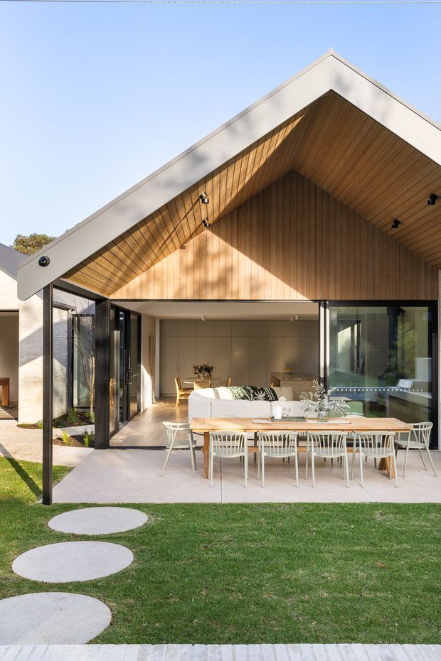 Exterior view of a contemporary gable-roof home opening onto a covered outdoor dining terrace. The facade combines pale rendered walls with a warm timber-lined gable soffit and wall, extensive black-framed glazing, and large hinged glass doors. A long timber dining table with white chairs sits on a concrete terrace beside a manicured lawn and circular stepping stones.