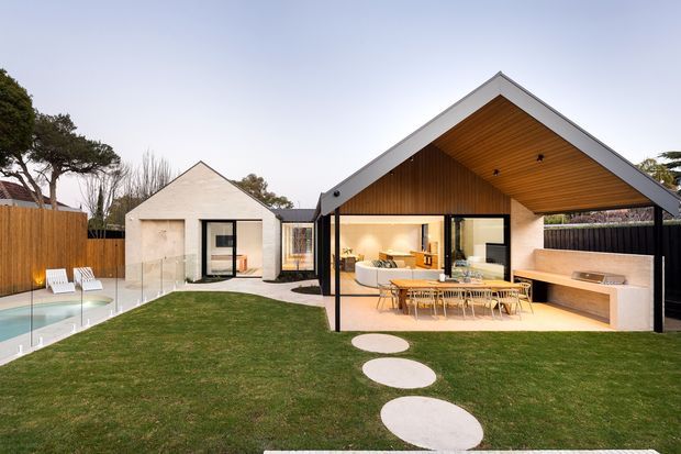 Contemporary single-storey residence viewed from the landscaped backyard, featuring a prominent pitched gable roof with a deep timber-lined eave over an open outdoor entertaining area. The main pavilion has extensive black-framed glazing, a built-in outdoor kitchen or barbecue bench, and a dining setting. A smaller matching gabled wing sits to the left beside a swimming pool, with timber fencing, lawn, and circular concrete stepping stones in the foreground.