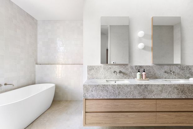 Bright contemporary bathroom with a freestanding white bathtub, large pale tiled walls, a double vanity with timber drawers and a mottled grey stone benchtop and splashback. Two rectangular mirrored cabinets and wall-mounted globe lights sit above the vanity.
