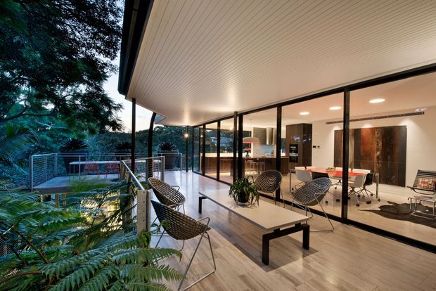 A contemporary indoor-outdoor living and dining space opening onto a landscaped elevated terrace. Floor-to-ceiling glazing connects the bright interior with the covered outdoor lounge, which features woven chairs, a low coffee table, timber-look flooring, and a slim metal balustrade. The interior includes a dining table, modern kitchen cabinetry, polished concrete-look or light stone flooring, recessed lighting, and dark-framed glazing. Lush vegetation surrounds the terrace.