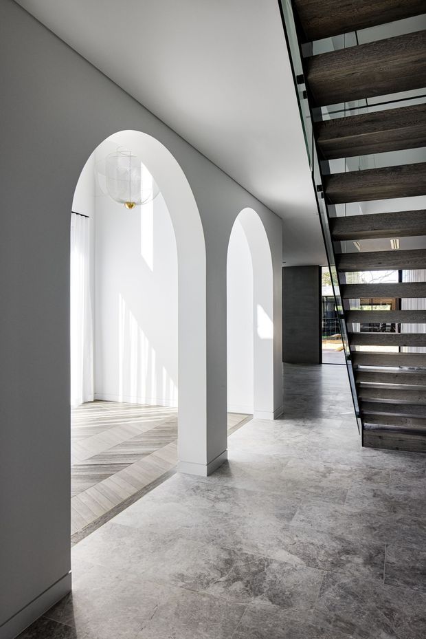 A refined contemporary interior hallway featuring tall white arched openings, pale grey walls, large-format grey stone flooring, and a dramatic open-riser staircase with dark timber treads and slim black supports. Strong daylight enters through the arched glazing, creating a bright, sculptural atmosphere. A pendant light hangs within the arched space.