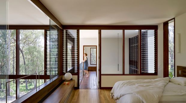 A bright modern bedroom with a low-profile bed, warm timber flooring and built-in timber joinery. Extensive floor-to-ceiling glazing and dark timber-framed louvred windows overlook a leafy bush setting. A glazed internal partition and corridor connect the bedroom to adjoining spaces, with strong horizontal and vertical lines throughout.