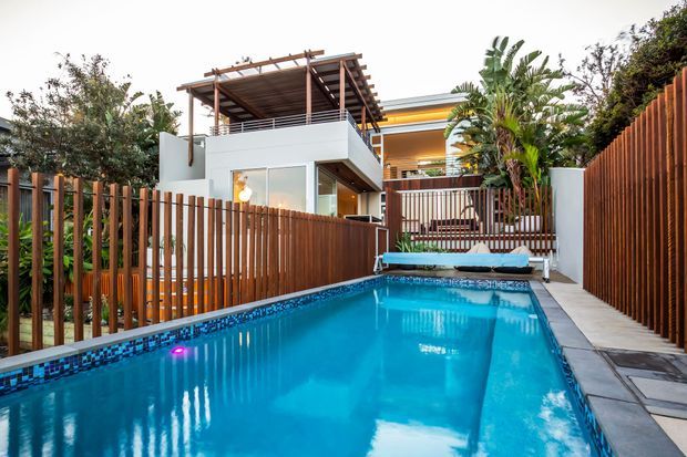 A modern multi-level residential house viewed from a narrow backyard swimming pool. The scene features a vivid blue tiled pool, timber slat privacy fencing, concrete or stone paving, tropical planting, and a contemporary white facade with extensive glazing. An upper-level rooftop or terrace structure with a pergola and timber screening is visible above the pool area.