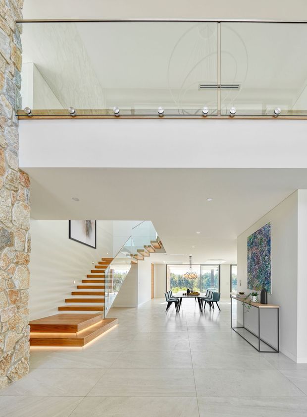 Bright contemporary open-plan interior viewed from an entry or hallway, featuring a floating timber staircase with glass balustrade, a double-height void with an upper-level glass balustrade, a large-format pale tiled floor, exposed stone wall, and a dining area beyond. Minimal white walls and ceilings are complemented by timber stair treads, black-framed furniture, and extensive glazing.