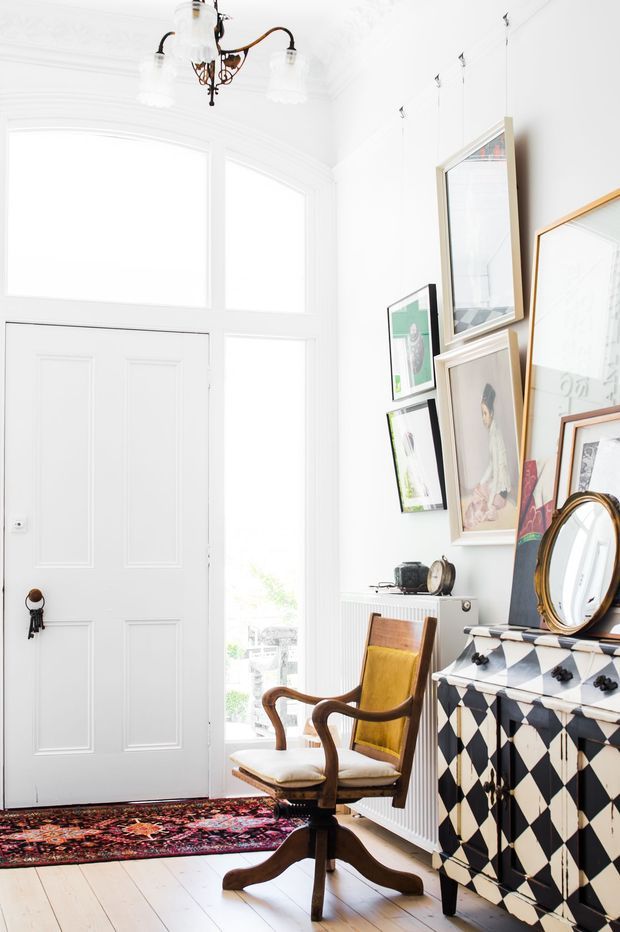 Bright, eclectic residential entryway or hallway with a tall white paneled door, large arched-top glazing, white walls and pale timber floorboards. A vintage chandelier hangs overhead, while a patterned Persian-style rug, mid-century wooden armchairs, gallery wall, ornate mirror and bold black-and-white diamond-patterned sideboard create a layered, characterful interior.