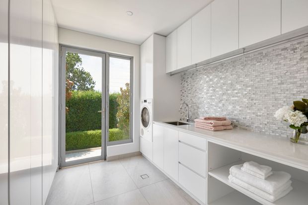 Bright, compact contemporary laundry with white floor-to-ceiling cabinetry, integrated front-loading washing machine, pale tiled flooring, a light stone-look benchtop, and a small stainless-steel sink. A silvery mosaic tile splashback runs along the work area, while a full-height glazed door opens to a clipped garden. Open shelving displays neatly folded towels, with soft pink towels and white flowers adding subtle colour.