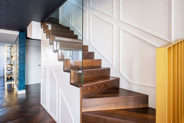 Interior view of a contemporary stairway with dark timber treads and a frameless glass balustrade. The stair wall features white decorative panel moulding, while a dark ceiling and patterned blue accent wall are visible in the adjoining hallway. A warm timber slatted element frames the right edge.