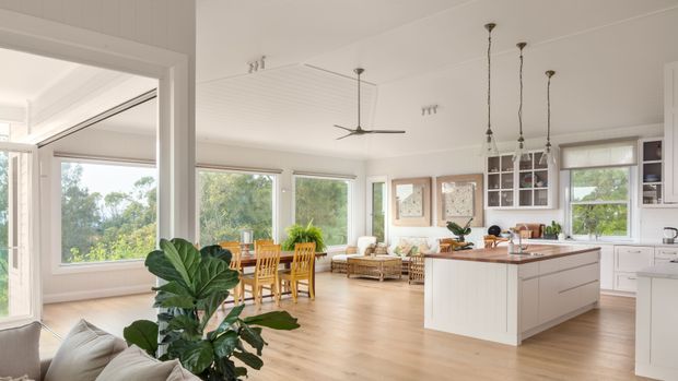 Bright, open-plan residential interior combining a kitchen, dining area and living space. The room features light timber flooring, white cabinetry and walls, a large central island with a timber benchtop, extensive windows, a dining table, lounge seating, pendant lights and a ceiling fan. Lush greenery is visible outside, contributing to an airy, relaxed atmosphere.