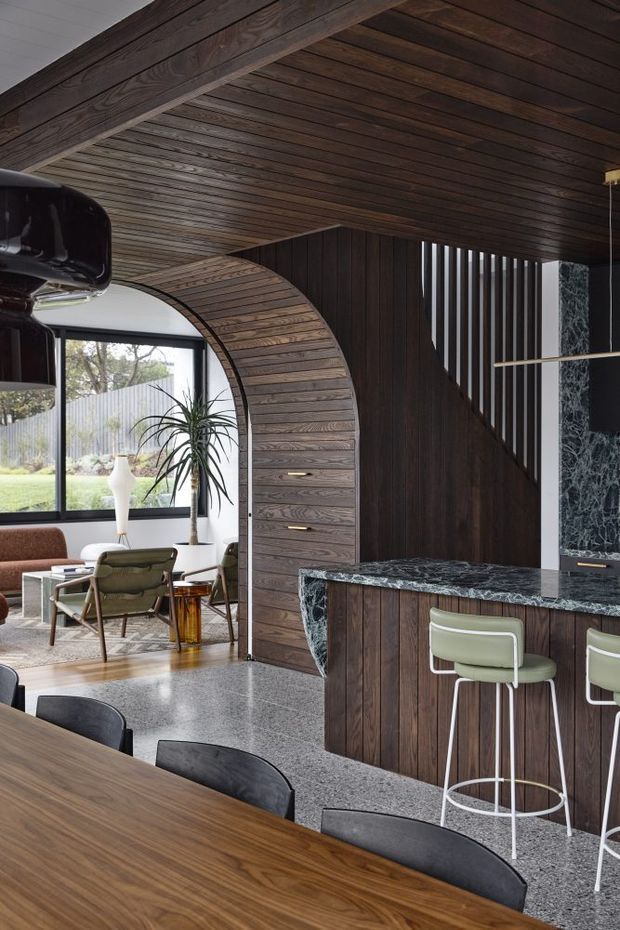 A sophisticated open-plan interior combining a dining area, kitchen and living space. Dark timber boards wrap the ceiling, structural beam and curved arched wall, creating a strong architectural feature. The kitchen has dark timber cabinetry, a veined dark stone benchtop, a raised peninsula with pale green bar stools, and integrated lighting. A terrazzo-like grey floor, timber dining table, black dining chairs, and mid-century-inspired lounge furniture complete the space. Large black-framed glazing opens the living area toward a garden.