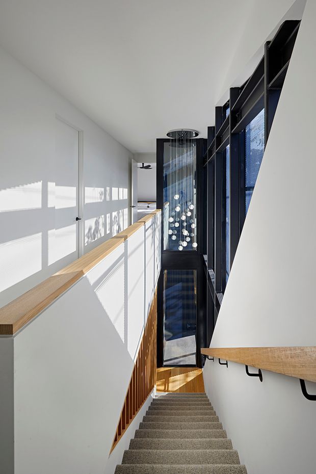 A bright, contemporary stairway and landing viewed from above. The stair descends through a narrow double-height void, with grey carpeted steps, white walls, light timber handrails, black metal supports and balustrades, and a dramatic vertical glazed feature with hanging spherical lights. Strong sunlight casts crisp blind patterns across the walls.