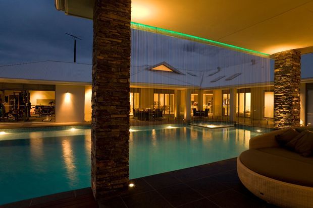 Dusk view of a sophisticated courtyard-style residence centred around a large illuminated swimming pool. Stone-clad structural columns frame the scene, while glazed living spaces open directly onto the pool deck. The building has a contemporary low-profile form with corrugated metal cladding, warm interior lighting, and a covered outdoor lounge area with rounded seating.
