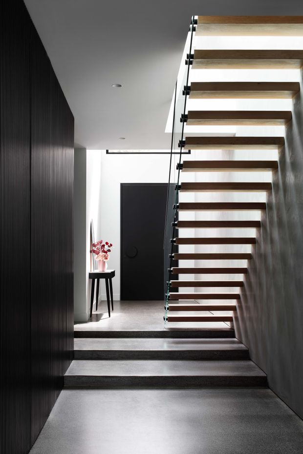 A contemporary interior stairway with floating dark timber treads, clear glass balustrades, and slim dark metal fittings. The stairs rise through a bright stairwell beside a textured dark wall, with polished grey flooring and several broad steps at the lower level. A matte black door and a small console table with flowers are visible in the background. A skylight or roof opening brings daylight into the vertical circulation space.