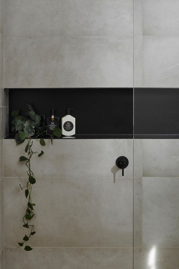 Close-up of a contemporary bathroom shower wall clad in large-format pale grey concrete-look tiles. A recessed horizontal black niche holds two pump bottles and a trailing green plant, with a minimalist matte-black shower control below. The composition is restrained, modern, and monochromatic with natural greenery as a small accent.
