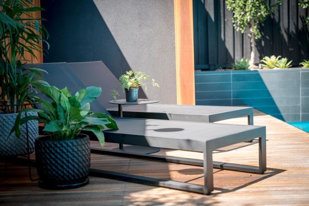 A sunlit outdoor poolside deck with two low-profile grey lounge tables, potted tropical plants, dark rendered walls, timber decking, and blue tiled pool edging. The scene has a clean contemporary design with strong geometric lines and warm natural light.