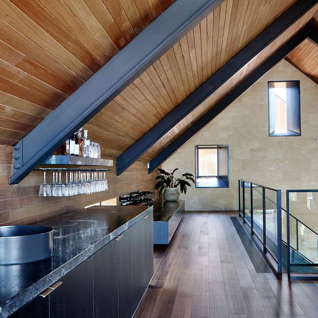 A sophisticated loft-like home bar beneath a steeply pitched timber-lined ceiling. The space features dark metal structural beams, a long dark stone bar counter with integrated cabinetry, suspended glassware storage, a small sink, and a stair or mezzanine balustrade with black metal and clear glass. Warm timber flooring and ceiling contrast with a pale stone feature wall and narrow windows.