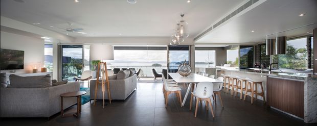 A spacious open-plan contemporary interior combining a living room, dining area, and kitchen. The room features floor-to-ceiling glazing with a water view, grey upholstered sofas, a timber dining table with mixed chairs, pendant lighting, dark tiled flooring, and a large kitchen island with timber cabinetry and light stone surfaces.
