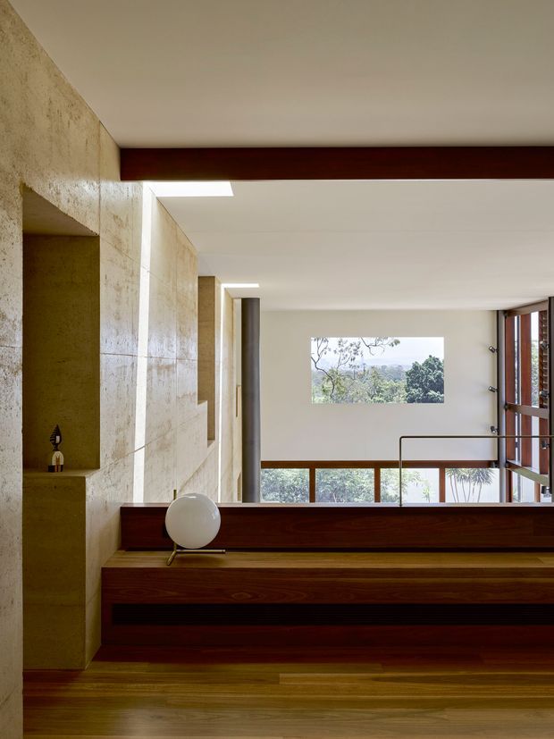 A refined modern interior with a warm timber floor and dark timber architectural beams, rails, and built-in joinery. Textured pale stone or plaster walls create recessed niches and openings, while slender metal columns, a minimalist stair or stepped platform, and large windows frame leafy views outside. The composition emphasizes strong horizontal and vertical lines, natural materials, and carefully controlled daylight.
