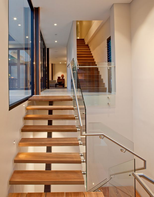 A contemporary residential stairway viewed from a lower level, featuring floating warm timber treads supported by a dark central spine, clear glass balustrades, and polished metal handrails. A second timber staircase rises in the background beside white walls and a tall glazed opening. Recessed ceiling lighting and abundant glazing create a bright, refined vertical circulation space.