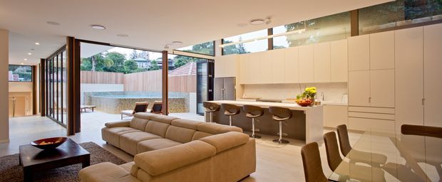 A bright, open-plan contemporary living space combining a living room, kitchen, and dining area. The room features extensive floor-to-ceiling glazing and timber-framed glass doors opening to a paved courtyard, a large cream sectional sofa, dark coffee table, pale timber flooring, streamlined light cabinetry, a kitchen island with stools, and a glass dining table. The interior has a clean, warm, spacious feel with strong indoor-outdoor connection.