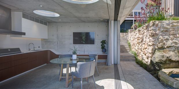 A contemporary open-plan kitchen and dining area with a restrained concrete-and-timber interior. The space features a long dark timber kitchen with integrated appliances, pale benchtops, a round glass dining table with upholstered chairs, exposed concrete ceilings and walls, circular skylights, and full-height glazed sliding doors opening to a stone-lined outdoor stair and courtyard area.