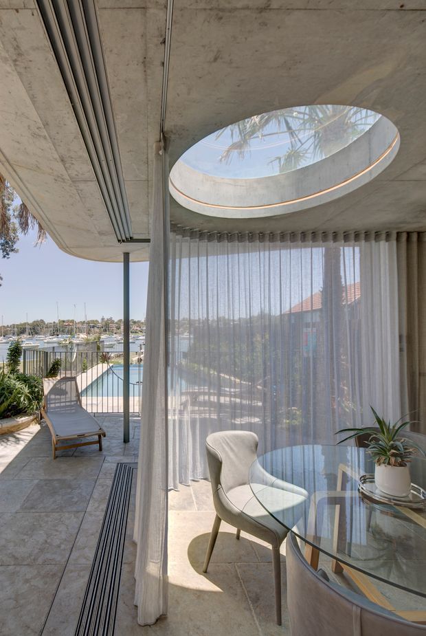 A bright, refined indoor-outdoor living space overlooking a marina. The image shows a curved glazed dining nook with sheer floor-to-ceiling curtains, a round glass dining table, upholstered dining chairs and a potted plant. An oval skylight punctures the exposed concrete ceiling, while a tiled terrace with a sun lounger, pool and waterfront beyond is visible to the left. The architecture features curved concrete forms, slender supports and extensive glazing.