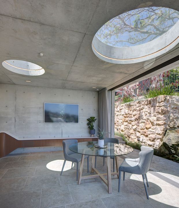 Contemporary dining area with a round glass dining table and upholstered chairs, set within a concrete-lined room. The space features large circular roof skylights, a wide glazed opening with retractable curtains, integrated timber-toned wall seating or cabinetry, stone floor tiles, and a landscaped rock wall visible outside.