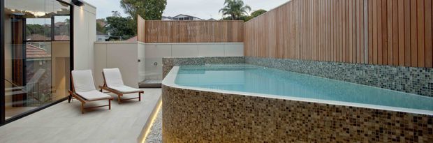 A contemporary private courtyard pool area with a raised tiled swimming pool, pale stone paving, timber privacy fencing, and frameless or dark-framed floor-to-ceiling glazing. Two white outdoor loungers sit beside the pool, while mosaic tile cladding wraps the pool walls and a narrow linear light accents the base.