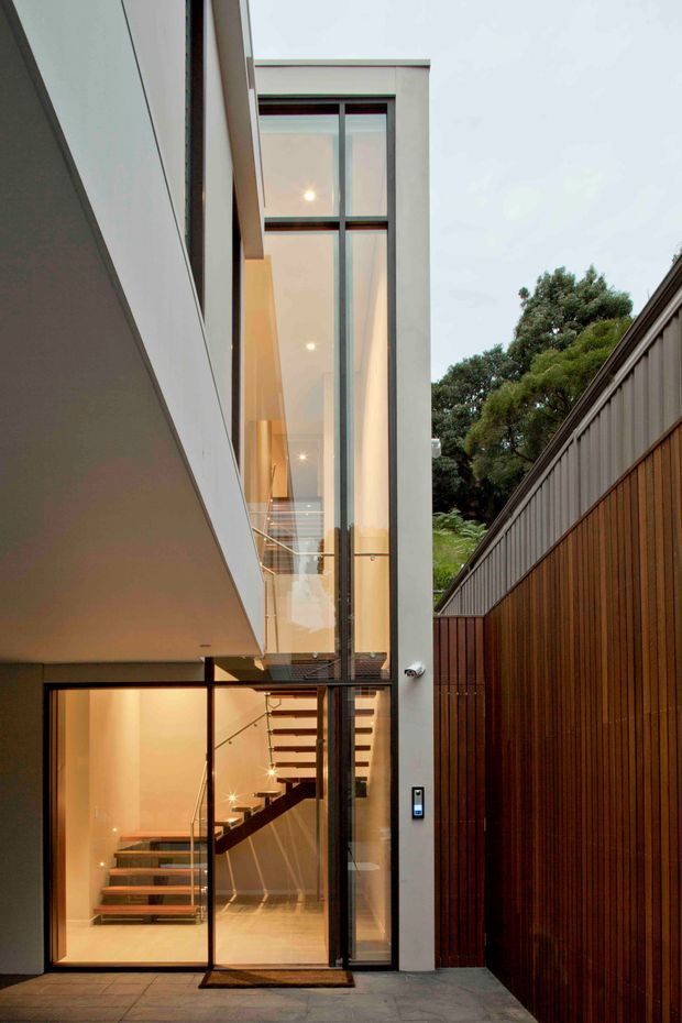 A narrow contemporary residential entry courtyard viewed from outside, featuring a tall glazed stairwell and entry door framed in dark metal, warm interior lighting, floating timber stair treads, and a sleek pale rendered facade. A vertical timber-clad boundary fence lines the right side, while mature trees are visible beyond. The composition emphasizes the dramatic multi-storey glazing, compact urban entry, and sculptural staircase.