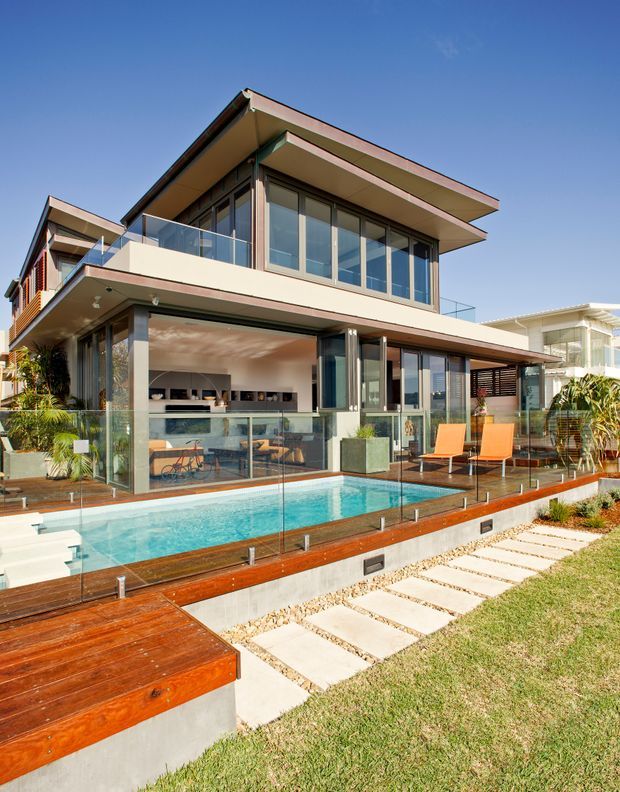 Exterior view of a modern multi-storey residence with broad projecting flat roof planes, extensive floor-to-ceiling glazing, glass balcony balustrades, and an open-plan ground floor. A rectangular swimming pool is bordered by timber decking and frameless glass safety fencing, with paved stepping stones and landscaped planting in the foreground.