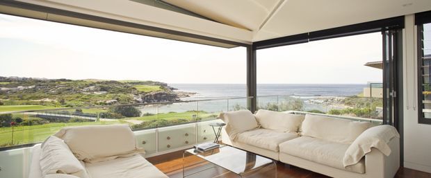 Bright contemporary living room with cream sofas and a glass coffee table, framed by expansive corner glazing and a glass balustrade overlooking a rugged coastal landscape and ocean. The room has a light neutral palette, dark window frames, and a distinctive angled ceiling detail.