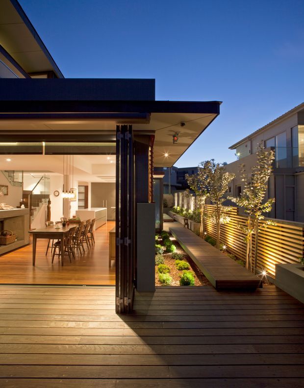 Dusk exterior view of a contemporary home opening onto a timber deck and landscaped side courtyard. Large retractable glazed doors reveal a warmly lit open-plan kitchen and dining area, while a narrow timber walkway, garden beds, and illuminated horizontal privacy screening extend along the side of the property. The home has strong horizontal roof planes, dark cladding or framing, and integrated outdoor lighting.