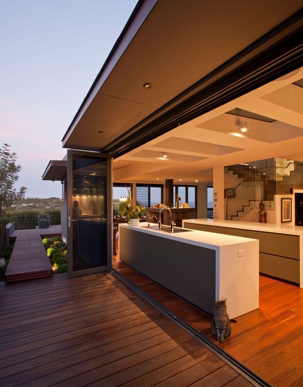 An open-plan contemporary kitchen opening directly onto a broad timber deck through large glazed folding or sliding doors. The kitchen features a long white island with a dark charcoal end panel, pale integrated cabinetry, stainless-steel tapware and appliances, warm timber flooring, and recessed ceiling lighting. A staircase and living area are visible beyond the kitchen, with landscaped surroundings and a distant waterfront or coastal outlook outside.